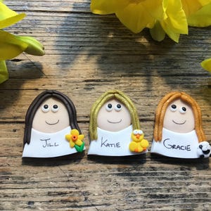 May include: Three handmade clay figurines with smiling faces, each with different hair colours and accessories. The figurines are named Jill, Katie, and Gracie. Jill has a flower, Katie has a chick, and Gracie has a sheep.