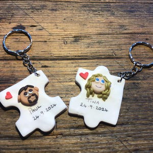 May include: Two white ceramic puzzle pieces with keyrings. Each piece features a cartoon face, a red heart, and a name with a date. The left piece says "Paul 24.9.2024" and the right piece says "Tina 24.9.2024".