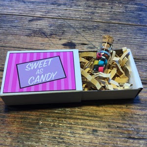 May include: A small white box with a pink and white striped lid. The lid has the text "SWEET AS CANDY" printed on it. The box is open and a small glass bottle filled with colorful candy is inside.