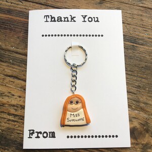 Op de afbeelding: Een witte kaart met de tekst "Thank You" en "From" erop gedrukt. Een zilveren sleutelhanger met een klein oranje figuurtje van een persoon met een bril en de tekst "Miss Sutcliffe" is aan de kaart bevestigd.