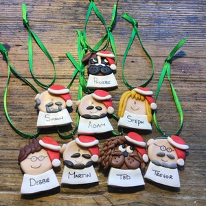 May include: Nine handmade Christmas ornaments featuring cartoon faces of people and a dog. Each ornament has a red Santa hat and a white name tag with a name written in black. The names are Simon, Adam, Steph, Debbie, Martin, Ted, Trevor, and Phoebe.