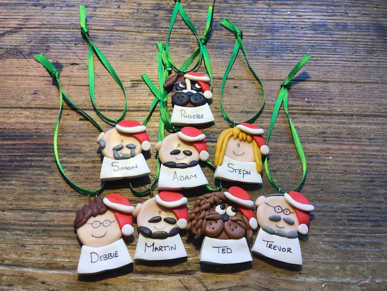 May include: Nine handmade Christmas ornaments featuring cartoon faces of people and a dog. Each ornament has a red Santa hat and a white name tag with a name written in black. The names are Simon, Adam, Steph, Debbie, Martin, Ted, Trevor, and Phoebe.