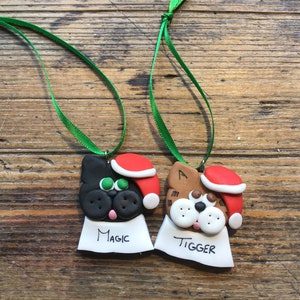 May include: Two handmade Christmas ornaments shaped like cats wearing Santa hats. The black cat ornament has green eyes and says "Magic" on the tag. The brown cat ornament has white eyes and says "Tigger" on the tag.