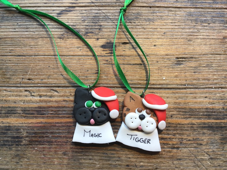 May include: Two handmade Christmas ornaments shaped like cats wearing Santa hats. The black cat ornament has green eyes and says "Magic" on the tag. The brown cat ornament has white eyes and says "Tigger" on the tag.