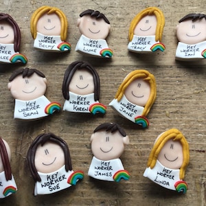 Op de afbeelding: Een verzameling van 12 kleurrijke sleutelwerknemer-badges met regenboogaccenten. Elke badge heeft een cartoongezicht met een naam en de tekst "Sleutelwerknemer" eronder.