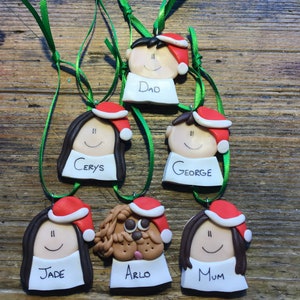 May include: Six personalized Christmas ornaments featuring cartoon faces with Santa hats. The ornaments are made of clay and have names written on them: Dad, Cerys, George, Jade, Arlo, and Mum.