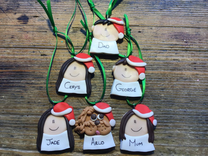 May include: Six personalized Christmas ornaments featuring cartoon faces with Santa hats. The ornaments are made of clay and have names written on them: Dad, Cerys, George, Jade, Arlo, and Mum.
