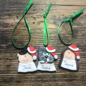 May include: Three handmade Christmas ornaments, each with a different face and name. The ornaments are made of clay and have a red Santa hat. The names are "Jack", "Pebble", and "Sophie".