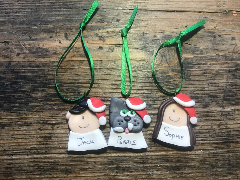 May include: Three handmade Christmas ornaments, each with a different face and name. The ornaments are made of clay and have a red Santa hat. The names are "Jack", "Pebble", and "Sophie".