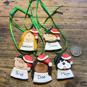 May include: Five handmade Christmas ornaments featuring cartoon faces with names written below. The ornaments are decorated with red Santa hats and green ribbon.