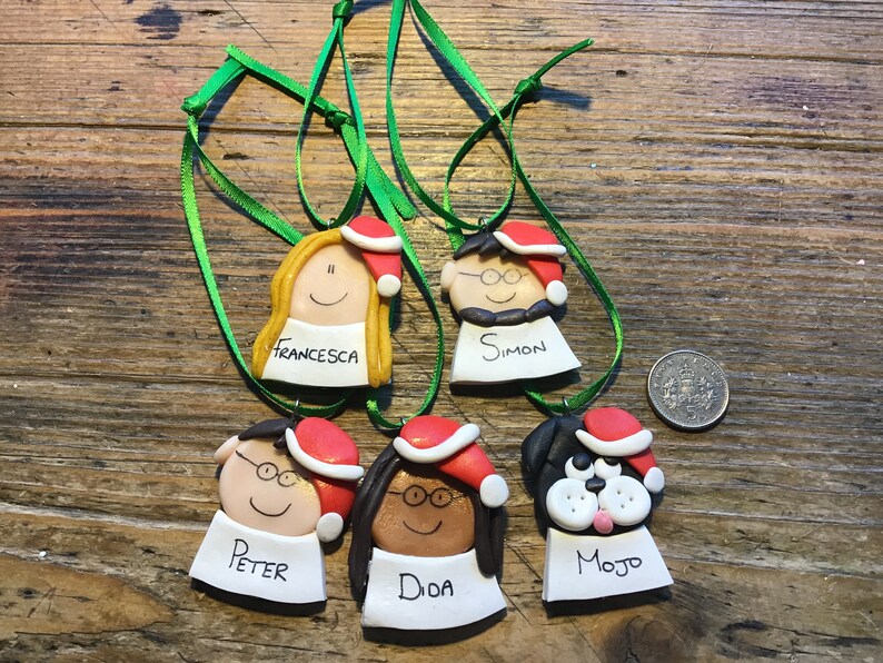 May include: Five handmade Christmas ornaments featuring cartoon faces with names written below. The ornaments are decorated with red Santa hats and green ribbon.