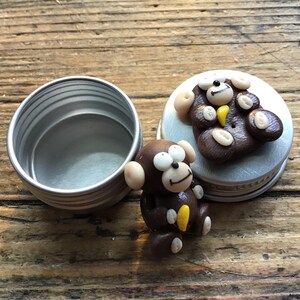 May include: A small, open, silver-colored tin next to a decorative monkey figurine and a lid with a monkey design. The monkey figurine is brown with tan ears and a yellow accent. The lid features a similar monkey design.