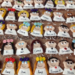May include: A collection of circular wine glass charms featuring miniature clay figures with diverse hair colours, floral headpieces, and white shirts. Each charm is personalised with a name written in black ink on the white base of the figure.