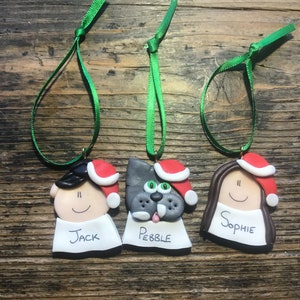 May include: Three handmade Christmas ornaments with green ribbon hangers. The ornaments are made of clay and feature a person with black hair, a dog with gray fur, and a person with brown hair. Each ornament has a red Santa hat and a white name tag. The names on the tags are Jack, Pebble, and Sophie.