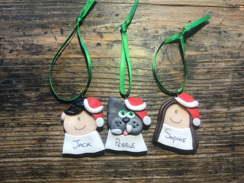May include: Three handmade Christmas ornaments with green ribbon hangers. The ornaments are made of clay and feature a person with black hair, a dog with gray fur, and a person with brown hair. Each ornament has a red Santa hat and a white name tag. The names on the tags are Jack, Pebble, and Sophie.