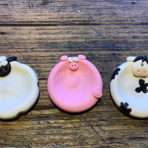 May include: Three small ceramic dishes shaped like farm animals. A white sheep, a pink pig, and a black and white cow.