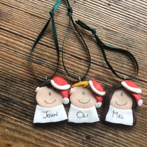 May include: Three personalized Christmas ornaments with red Santa hats, white faces, and black eyes. The ornaments are labeled "John", "Oli", and "Mel".