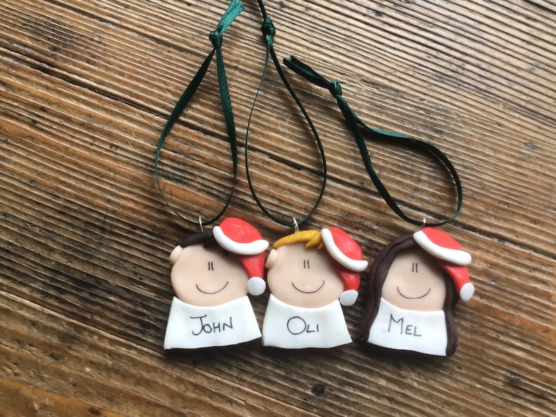 May include: Three personalized Christmas ornaments with red Santa hats, white faces, and black eyes. The ornaments are labeled "John", "Oli", and "Mel".