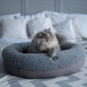 Puede incluir: Un gato gris con pelaje blanco alrededor de la cara y las patas est&aacute; acostado en una cama redonda gris para mascotas. La cama est&aacute; sobre una manta de patr&oacute;n gris y blanco.