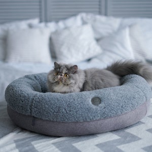 Puede incluir: Un gato gris con ojos amarillos se sienta en una cama redonda para mascotas de color gris sobre una manta blanca y gris. La cama tiene un forro suave y peludo.