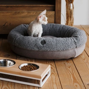 Puede incluir: Una estaci&oacute;n de comida para mascotas de madera con tres cuencos de acero inoxidable. La estaci&oacute;n tiene un marco de metal blanco y est&aacute; elevada del suelo. Un gato est&aacute; sentado en una cama para mascotas gris en el fondo.