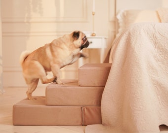 May include: A light brown pug dog climbing up a set of four beige pet steps. The steps are in front of a white bed with a white and beige quilt.