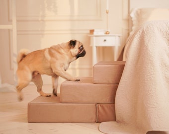 May include: A light brown pug dog is climbing up a set of three beige pet steps. The steps are in front of a white bed with a white and beige patterned comforter.