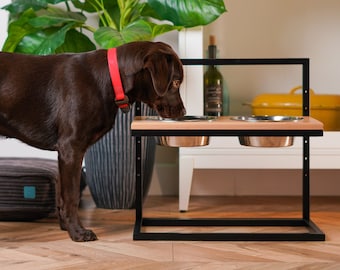 May include: A brown dog eating from a raised double bowl pet feeder. The feeder is made of black metal and wood with two stainless steel bowls. The dog is standing on a wood floor.