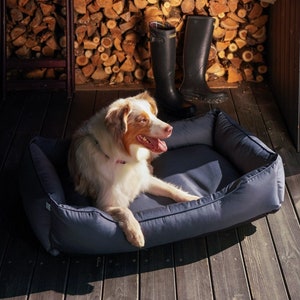 May include: A brown and white dog laying in a dark blue dog bed. The dog bed is on a wooden floor. There are two pairs of black rain boots in the background.