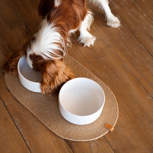 May include: A brown and white dog eating from a white ceramic bowl on a brown woven mat. There is a second empty white ceramic bowl on the mat.