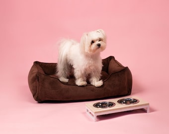 Op de afbeelding: Een kleine witte hond die op een bruin corduroy hondenbed zit. De hond kijkt naar de camera. Er staan twee roestvrijstalen hondenbakken op een witte houten standaard voor het bed.