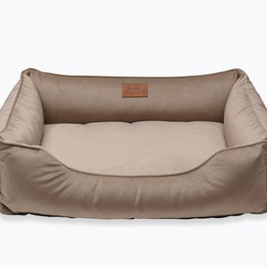 May include: A rectangular dog bed in a neutral beige color. The bed has a plush, cushioned design with raised sides and a lower front. A small, rectangular patch with text is centered on the back of the bed.