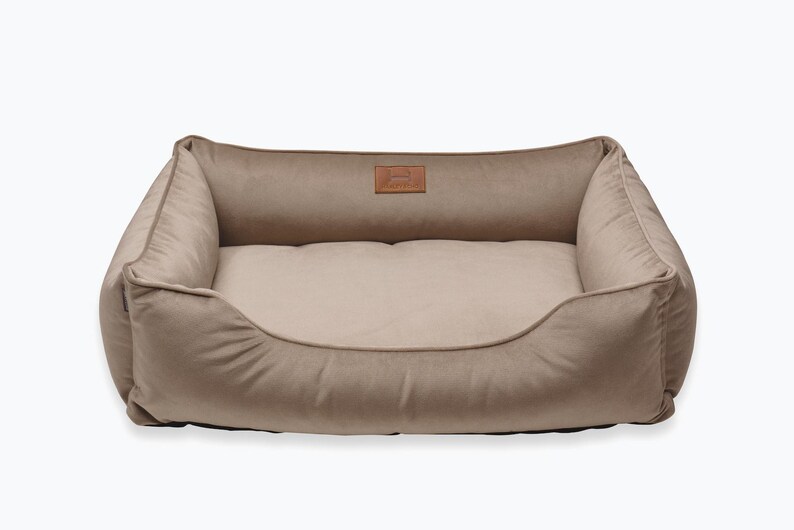 May include: A rectangular dog bed in a neutral beige color. The bed has a plush, cushioned design with raised sides and a lower front. A small, rectangular patch with text is centered on the back of the bed.