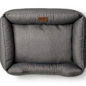 May include: Gray rectangular pet bed with raised sides and a plush interior. The bed features a brown leatherette tag with a logo. The bed is designed for comfort and support, perfect for a dog or cat to rest.