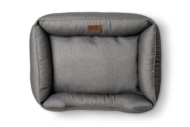May include: Gray rectangular pet bed with raised sides and a plush interior. The bed features a brown leatherette tag with a logo. The bed is designed for comfort and support, perfect for a dog or cat to rest.