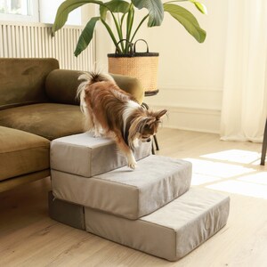 May include: A small brown and white dog is climbing up a set of three grey fabric steps. The steps are in a living room with a green sofa and a potted plant in the background.