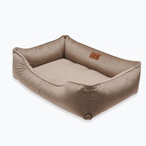 May include: A rectangular dog bed in a neutral beige color. The bed has a soft, padded interior and raised sides for comfort. A small, brown leather patch with a logo is sewn on the side. The bed is designed for pets.