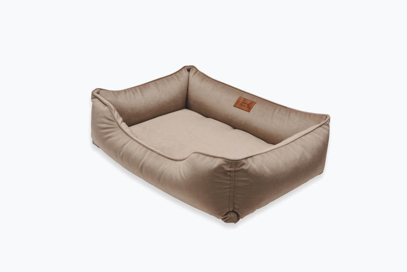 May include: A rectangular dog bed in a neutral beige color. The bed has a soft, padded interior and raised sides for comfort. A small, brown leather patch with a logo is sewn on the side. The bed is designed for pets.