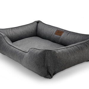 May include: A rectangular dog bed in a dark gray fabric. The bed has raised sides and a lower front, with a lighter gray interior. A small, brown leather patch with a logo is sewn on the side.