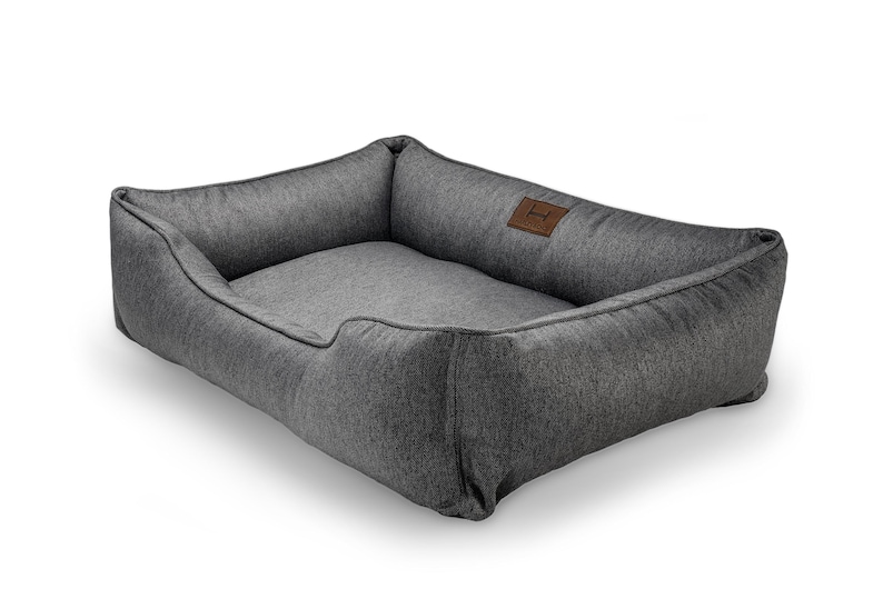 May include: A rectangular dog bed in a dark gray fabric. The bed has raised sides and a lower front, with a lighter gray interior. A small, brown leather patch with a logo is sewn on the side.