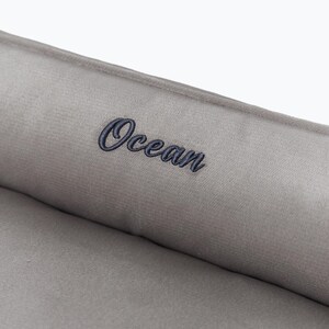 May include: Close-up of a gray, plush fabric with the word "Ocean" embroidered in a dark blue script. The fabric appears soft and textured, suggesting a comfortable item.