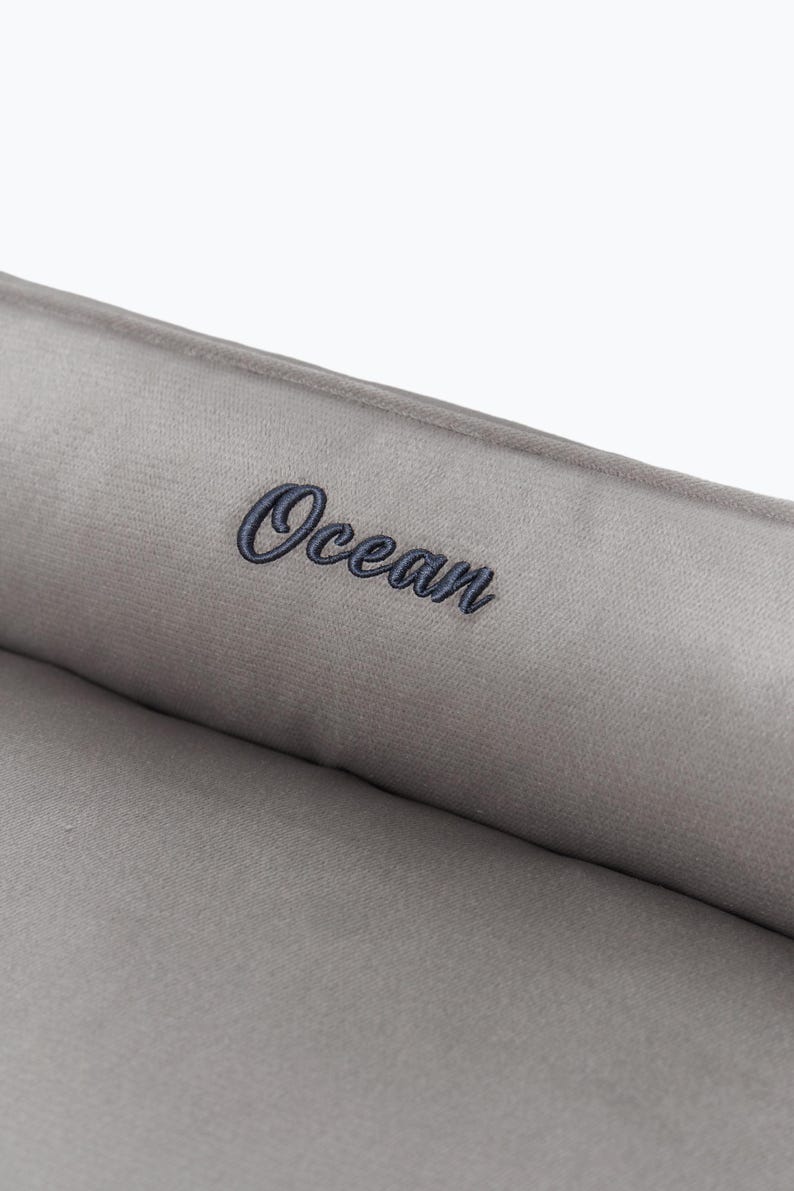 May include: Close-up of a gray, plush fabric with the word "Ocean" embroidered in a dark blue script. The fabric appears soft and textured, suggesting a comfortable item.