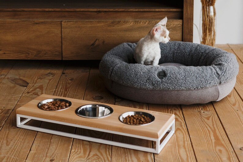 Puede incluir: Una estaci&oacute;n de comida para mascotas de madera con tres cuencos de acero inoxidable. La estaci&oacute;n tiene un marco de metal blanco y est&aacute; elevada del suelo. Un gato est&aacute; sentado en una cama para mascotas gris en el fondo.