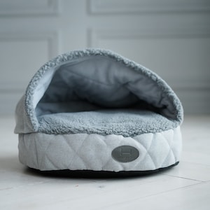Puede incluir: Una cama para perros acolchada gris y blanca con un forro suave y mullido. La cama tiene una abertura con capucha para mayor comodidad y seguridad.