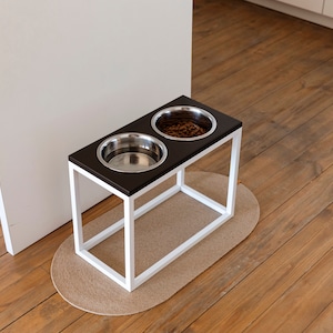 May include: White elevated dog bowl stand with two stainless steel bowls. One bowl is filled with dog food. The stand is on a light brown wood floor with a beige oval rug.