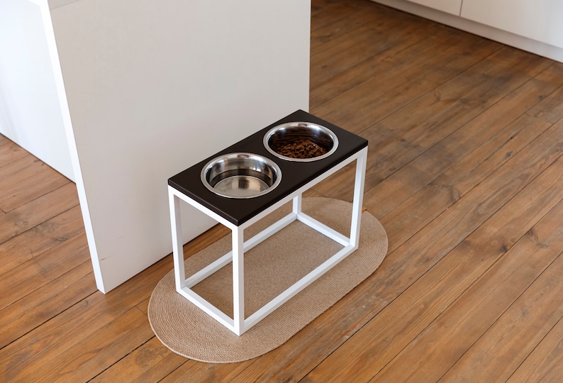 May include: White elevated dog bowl stand with two stainless steel bowls. One bowl is filled with dog food. The stand is on a light brown wood floor with a beige oval rug.