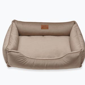May include: A beige rectangular pet bed with a plush, cushioned interior and raised sides. The bed features a small, rectangular leather patch with a logo on the headrest. The overall design is simple and comfortable.