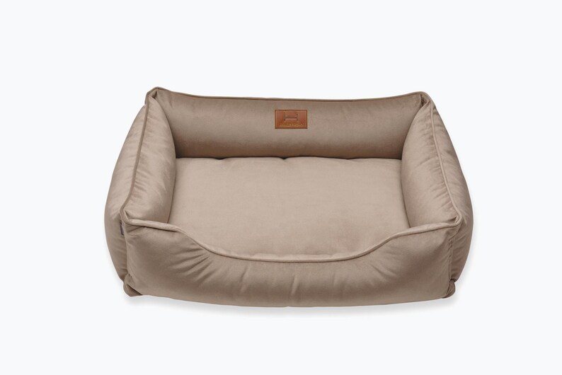 May include: A beige rectangular pet bed with a plush, cushioned interior and raised sides. The bed features a small, rectangular leather patch with a logo on the headrest. The overall design is simple and comfortable.