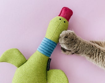 Puede incluir: Un juguete de peluche verde en forma de pato con un pico rojo y un cuello a rayas azul y blanco. Una pata de gato alcanza el juguete.