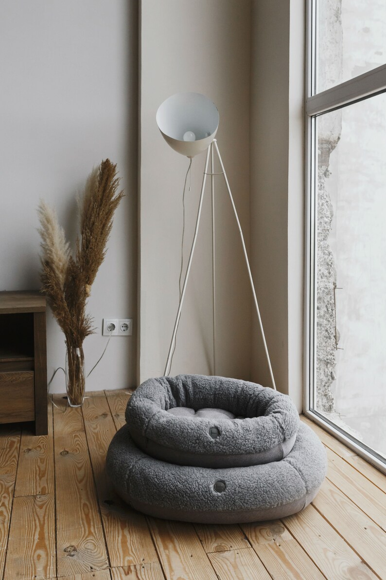 Puede incluir: Una cama redonda para mascotas de color gris con textura suave se encuentra en un suelo de madera. En el fondo se ve una l&aacute;mpara de pie blanca con un soporte de tr&iacute;pode.